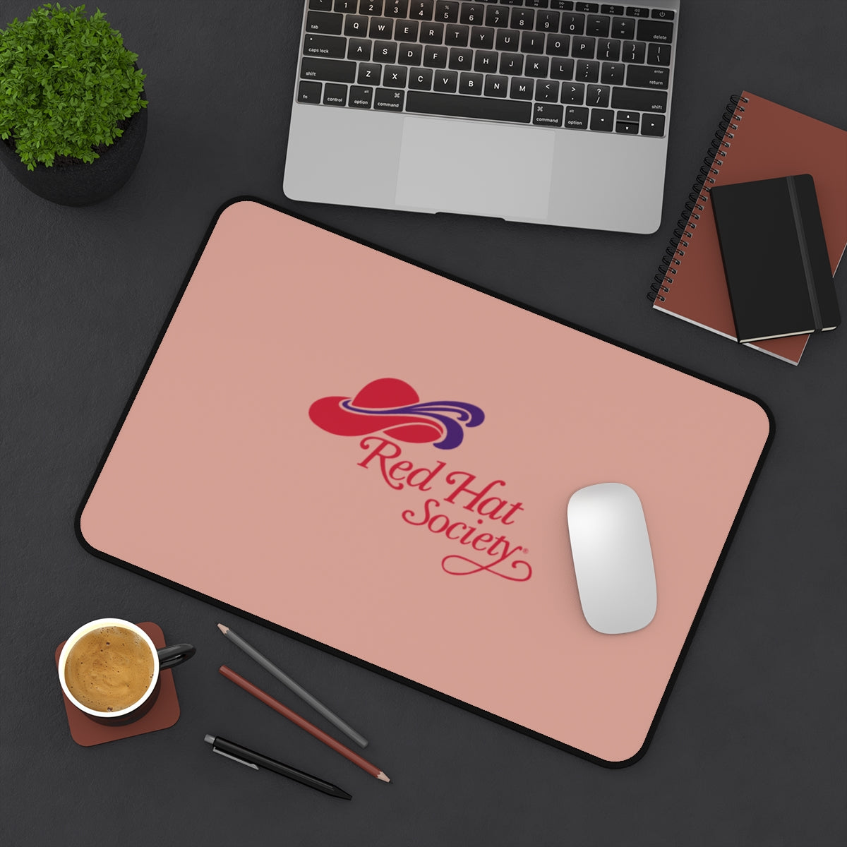 Large blush pink desk mat featuring the Red Hat Society logo in red and purple, styled on a modern workspace with a laptop, coffee and notebooks - perfect for adding personality and comfort to any desk.